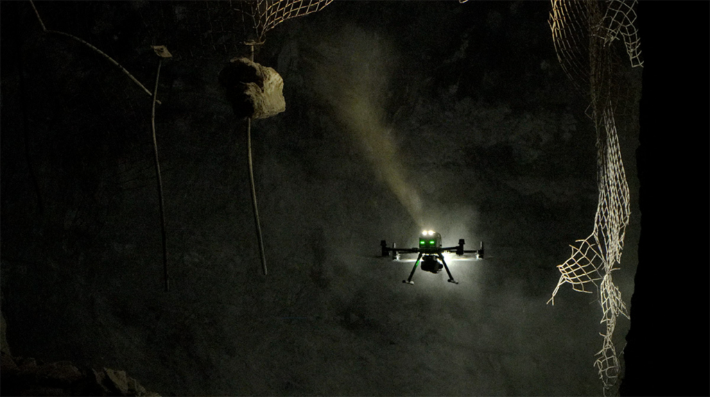 A drone with green lights flies in a dark cave, surrounded by rock walls, metal rods, and torn safety nets.