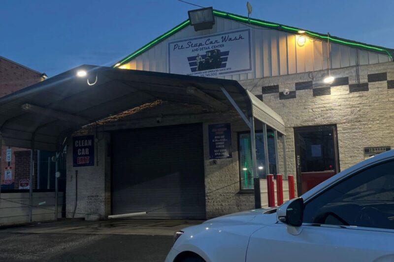 A closed car wash building at dusk, with a sign reading "Pit Stop Car Wash and Detail Center" above a shuttered garage door and a white car parked in front.