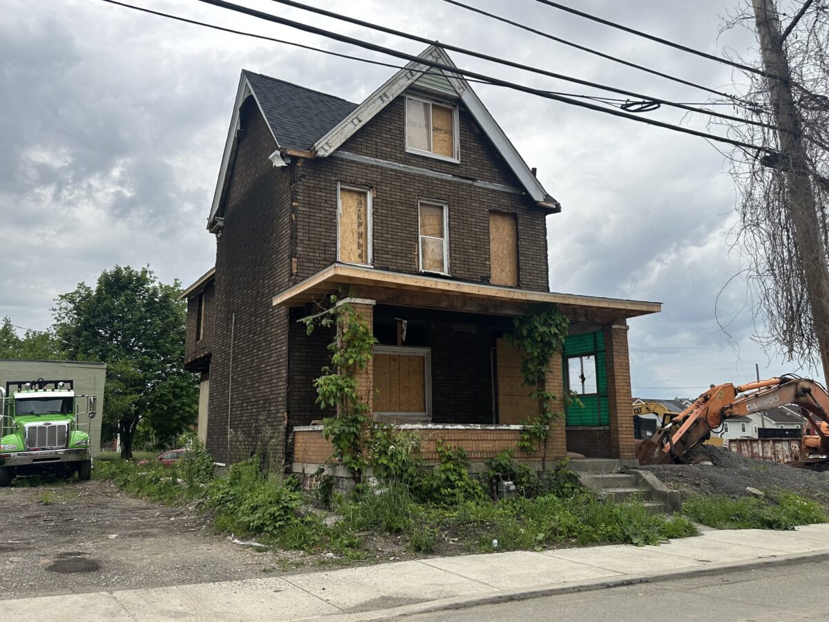 A boarded-up, two-story brick house with overgrown plants on the porch sits next to an excavator and a green truck under a cloudy sky.