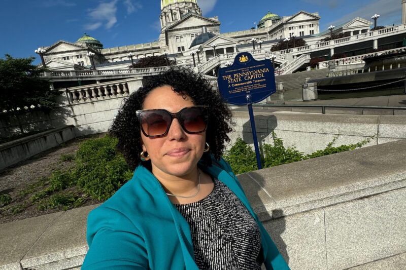 A person in sunglasses and a teal blazer takes a selfie in front of the Pennsylvania State Capitol building on a sunny day.