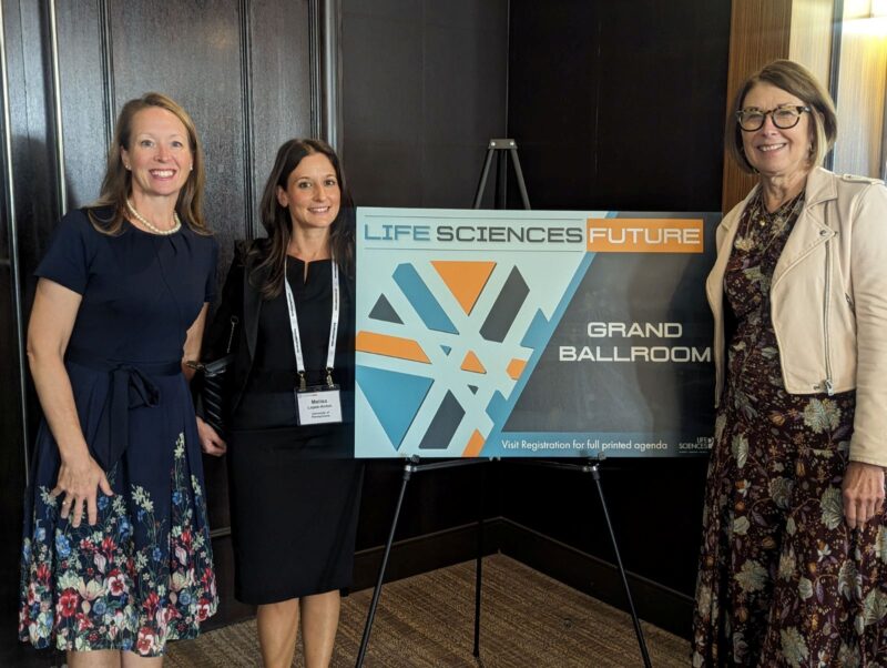 Three women stand beside a sign that reads "Life Sciences Future Grand Ballroom" at an indoor event.