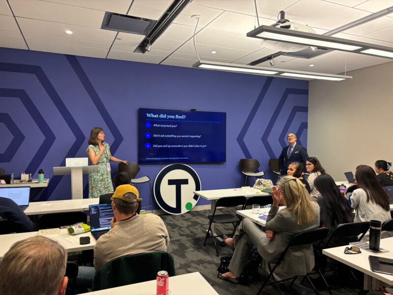 A white woman in a green dress stands next to a TV screen that reads "What did you find?" in front of a group of people sitting at white desks