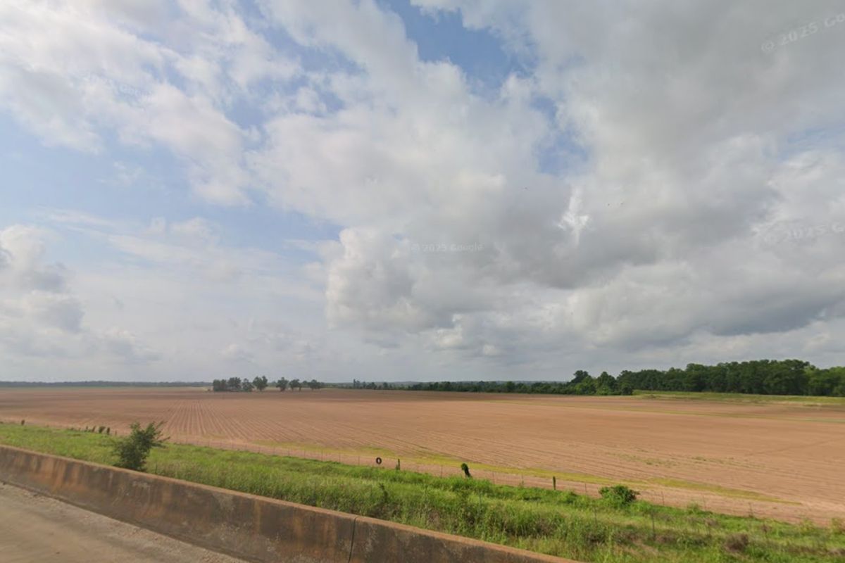A wide, flat, freshly plowed field stretches under a partly cloudy sky, bordered by trees and a low concrete barrier in the foreground.