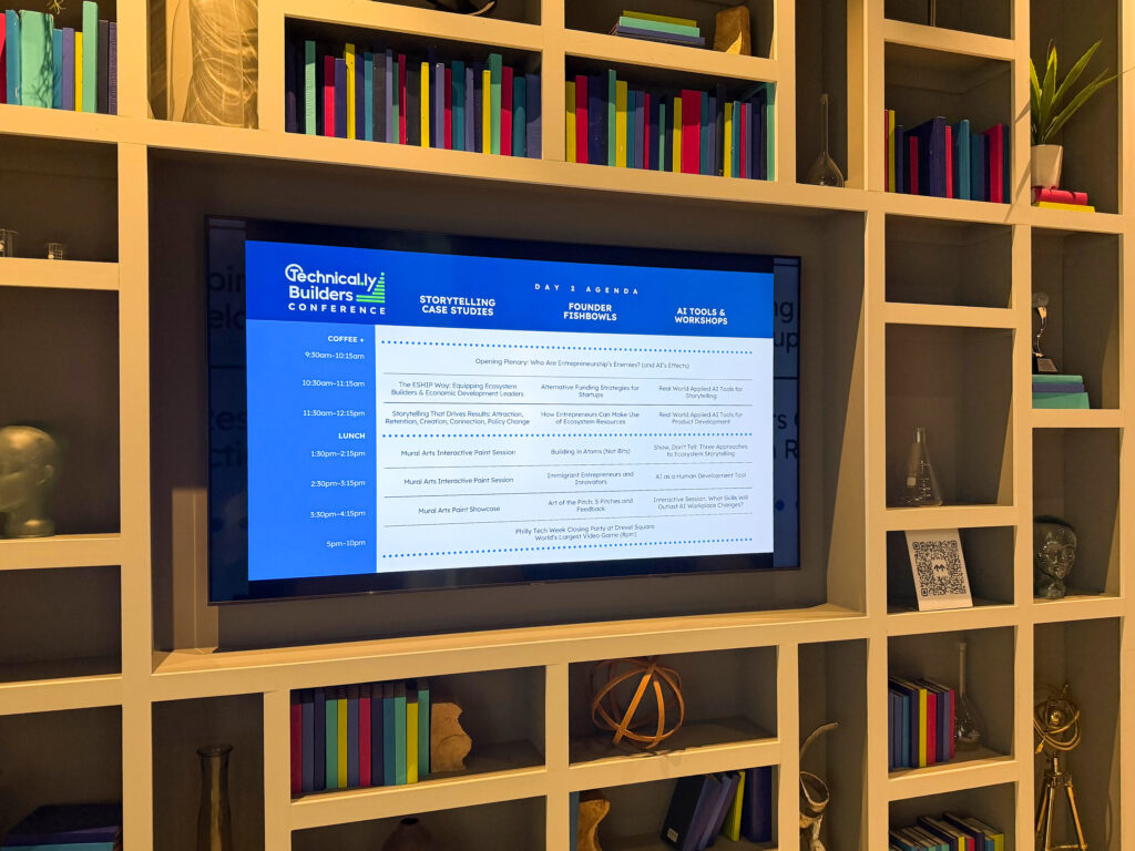 A TV screen displays a schedule for the Technically Builders Conference, surrounded by a bookshelf filled with books and decorative items.