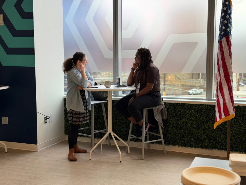 Two women sit at a high table in front of large windows, engaged in conversation. An American flag stands to the right, and geometric patterns decorate the walls.