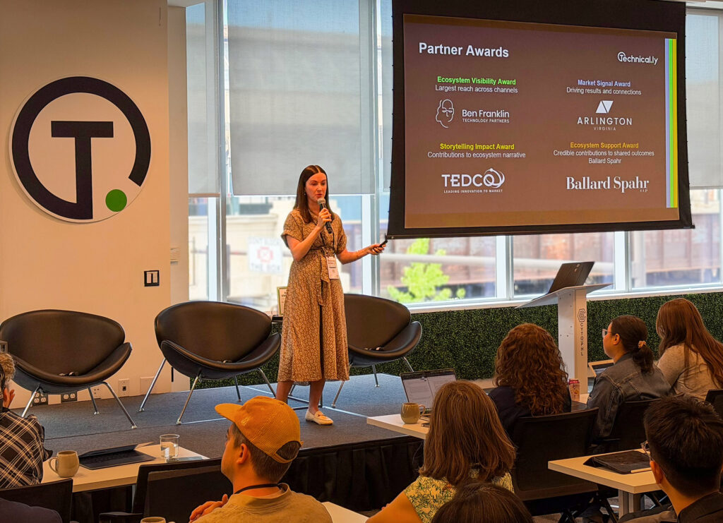 A woman speaks on stage with a microphone during a presentation. A slide behind her displays partner awards. Audience members are seated and listening.