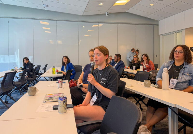 A young woman speaks into a microphone at a conference room meeting, with several people seated at tables listening and taking notes.