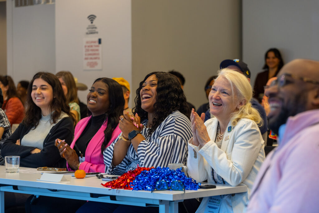 A group of people sit at tables, smiling and clapping during an indoor event or presentation. Pom-poms and drinks are on the table.