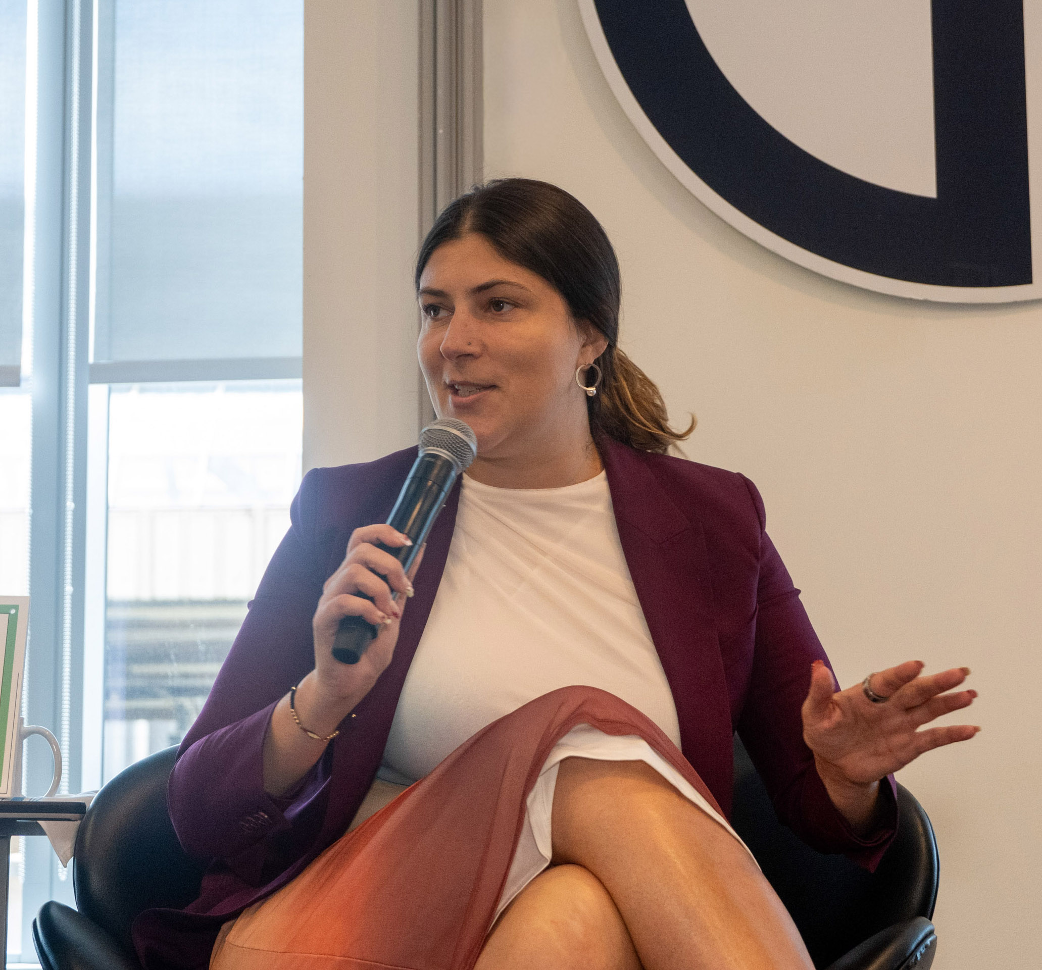 A woman in a maroon blazer and white dress sits in a chair, holding a microphone and gesturing with her hand, speaking during an indoor event.