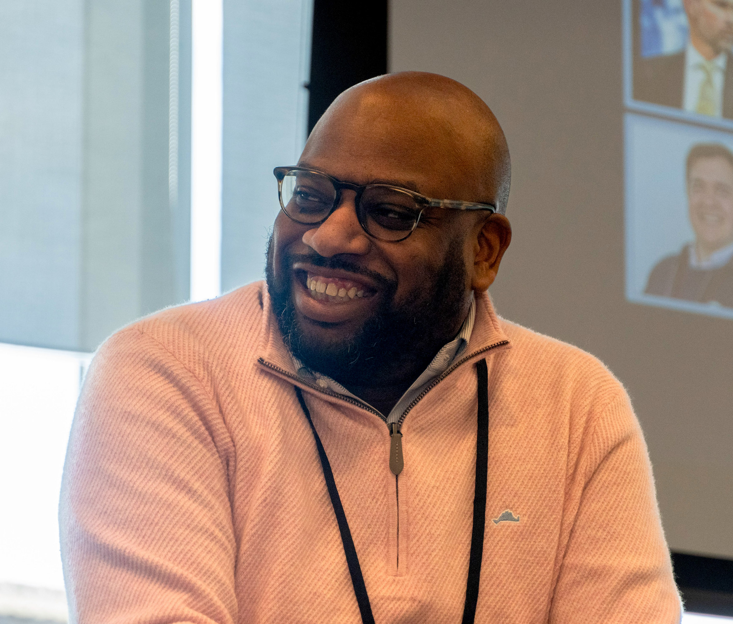 A man wearing glasses and a light pink sweater smiles while sitting indoors, with a projector screen and window in the background.