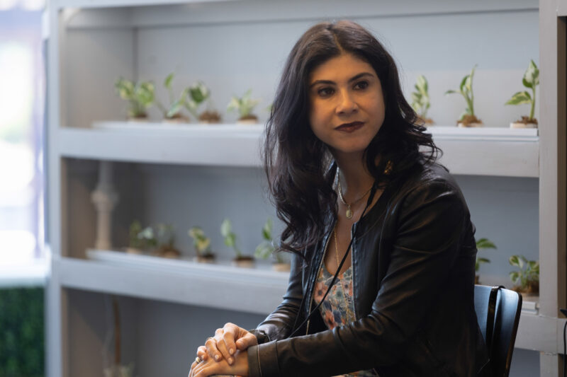 A woman with dark hair, wearing a black leather jacket, sits indoors near shelves with small potted plants.