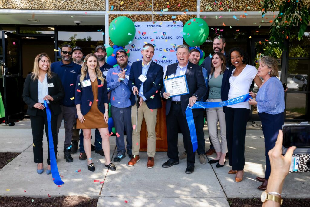 A group of people stand in front of a building at a ribbon-cutting ceremony, with one man holding a certificate and others smiling amid falling confetti and balloons.