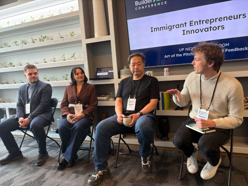Four panelists sit on chairs in front of a screen displaying "Immigrant Entrepreneurs & Innovators" at a conference, engaged in discussion.