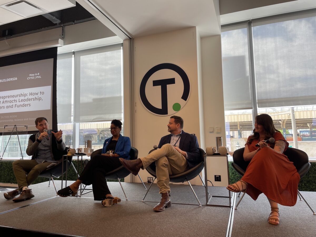 Four panelists sit on a stage in front of a screen, engaged in a discussion, with large windows and a round logo in the background.