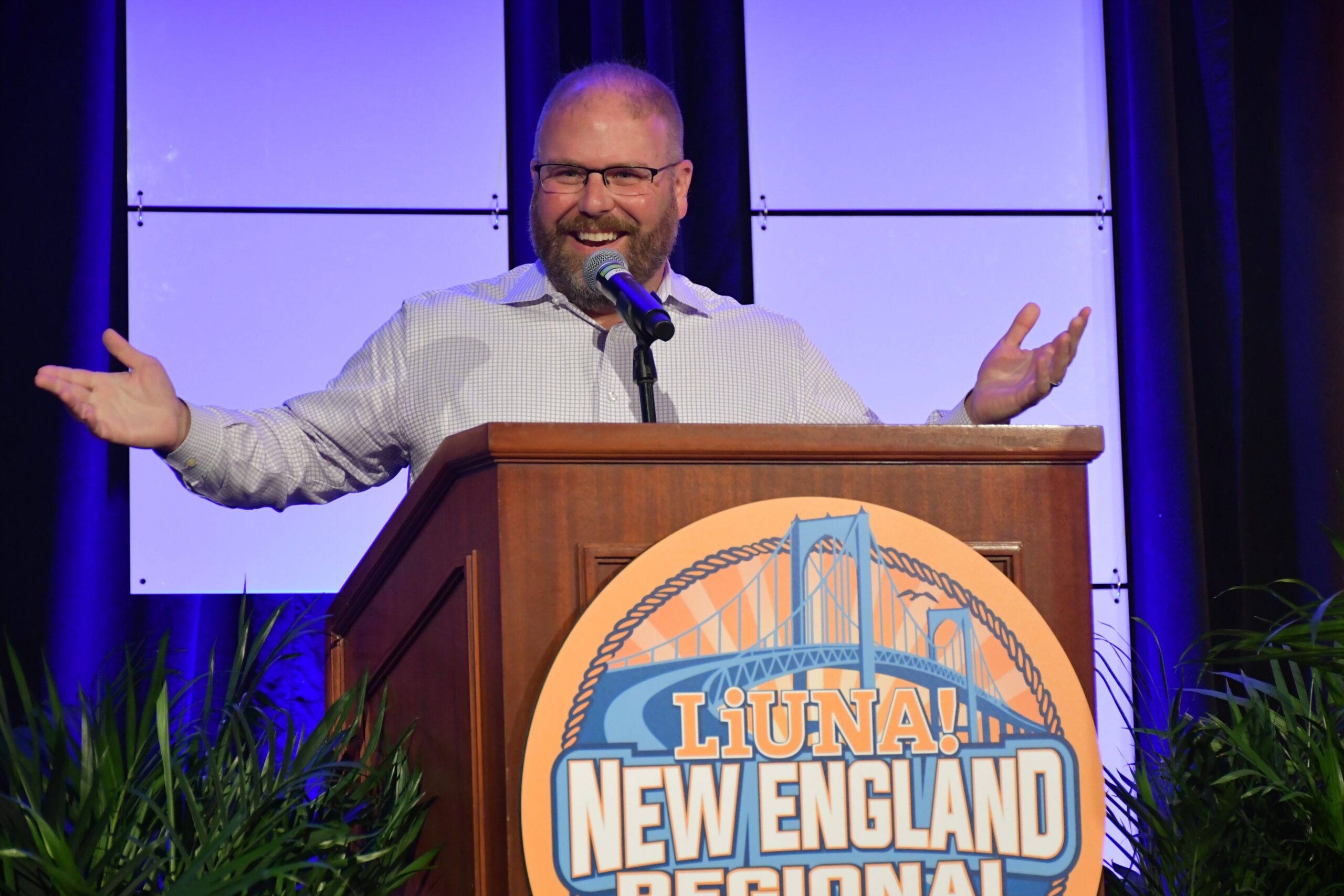 A man with a beard speaks at a podium with a LiUNA! New England Regional sign, smiling with arms open, against a blue-lit background.