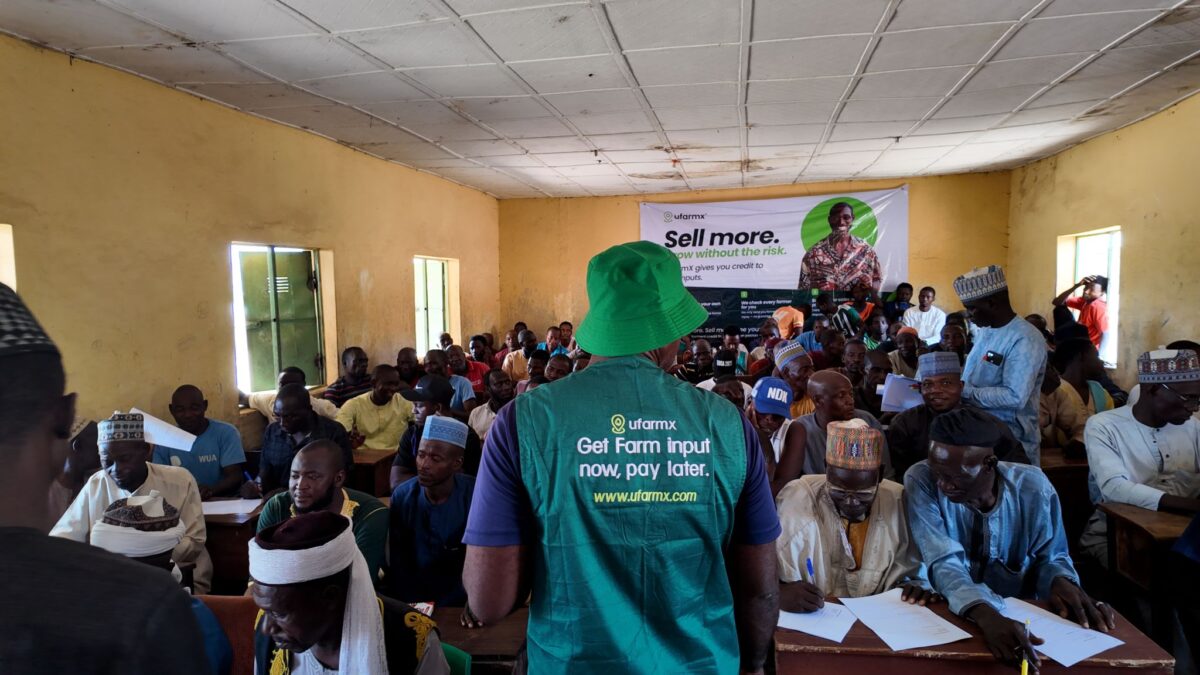 A Black man wears a green shirt that says "ufarmx" in the foreground. In the background, a room of Black men are filling out forms.