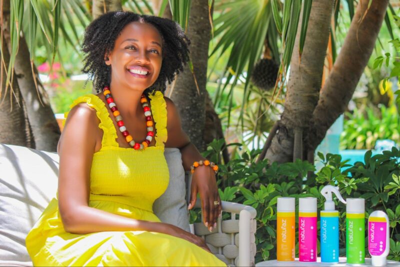 A woman in a yellow dress sits outdoors on a bench, smiling, with several colorful Naturaz hair product bottles displayed beside her.