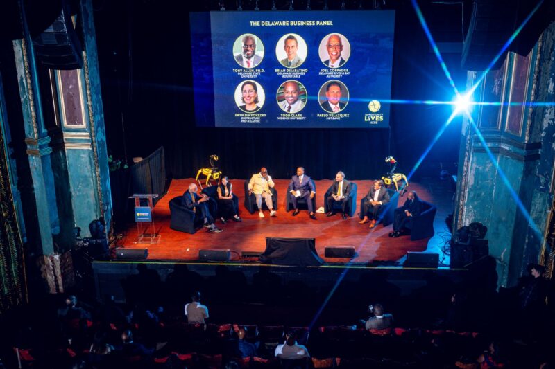 A panel of seven people sits on stage in front of an audience, with a large screen behind them displaying “The Delaware Business Panel” and panelist photos and names.