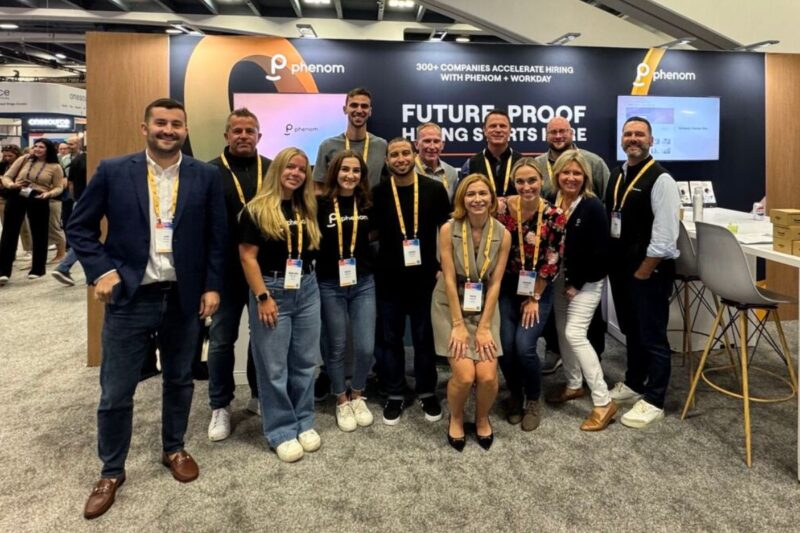 A group of people pose together and smile in front of a Phenom booth at a convention, with event badges around their necks and a conference display in the background.