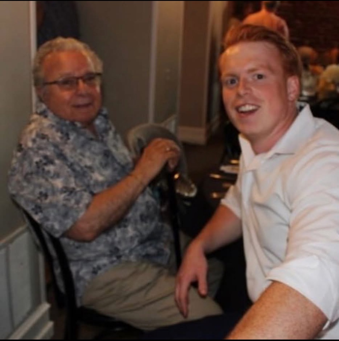 Two men sitting at a dining table, one older in a patterned shirt and glasses, the other younger in a white shirt, both looking toward the camera.