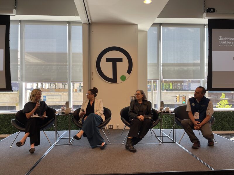 Four people sit on a stage in discussion, with two women and one man facing a moderator, under a large circular logo on the wall behind them.