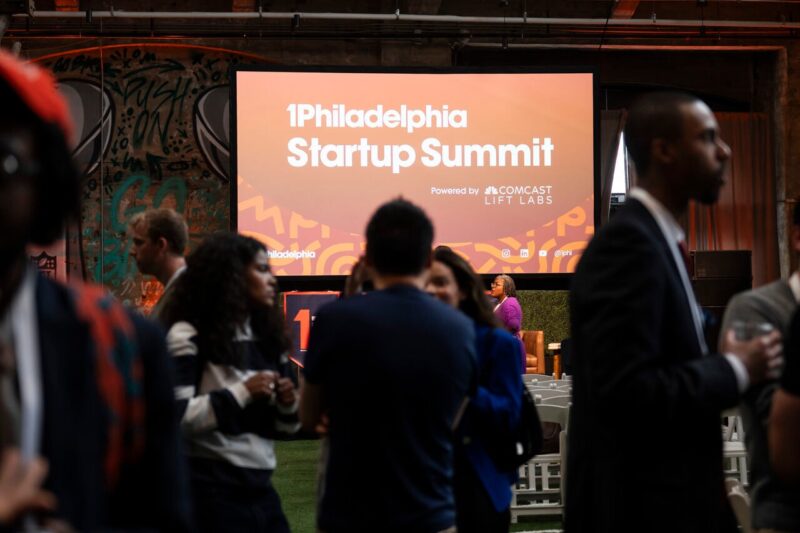 People gather in front of a large screen displaying "Philadelphia Startup Summit Powered by Comcast Lift Labs" at an indoor event space.