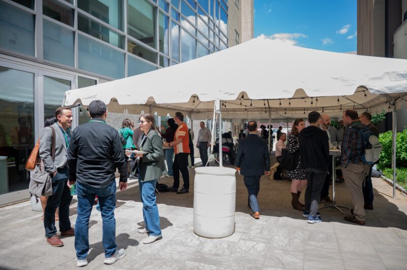 People gather and talk under a white outdoor tent set up beside a modern building on a sunny day.
