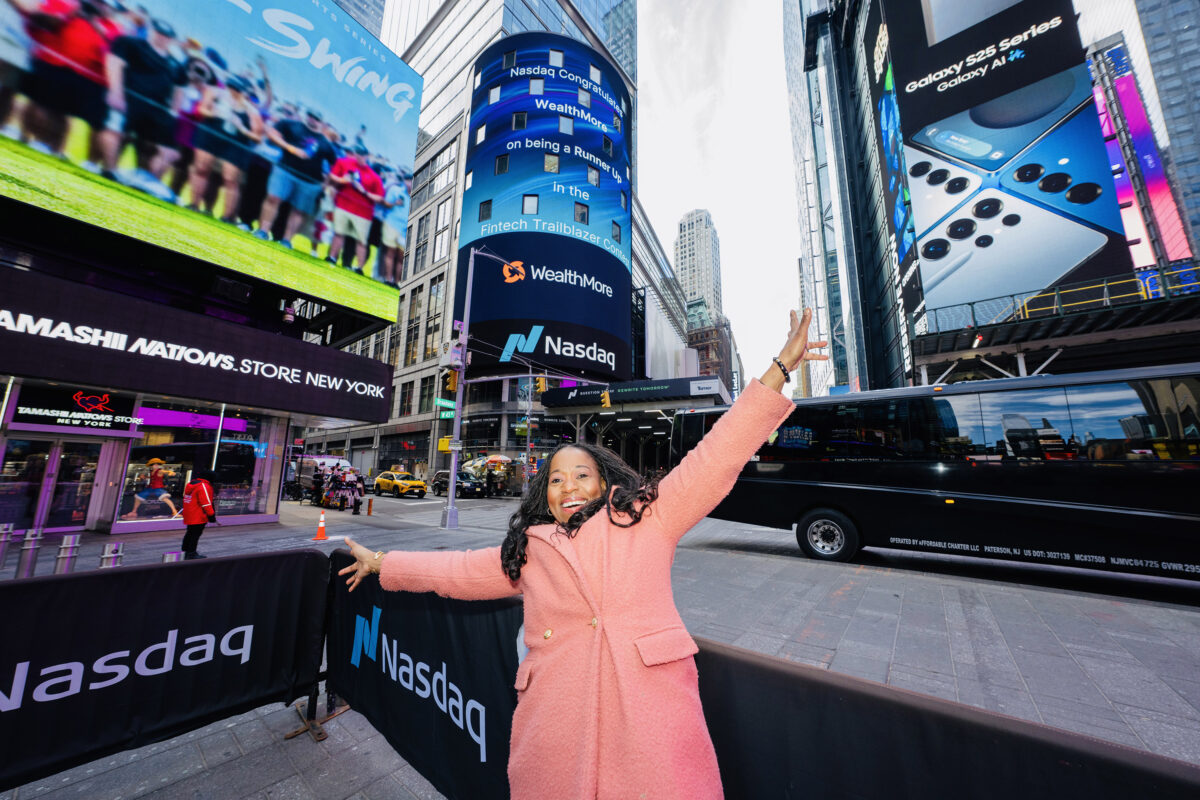 A woman in a pink coat smiles and raises her arms in Times Square, New York City, with Nasdaq and digital billboards in the background.