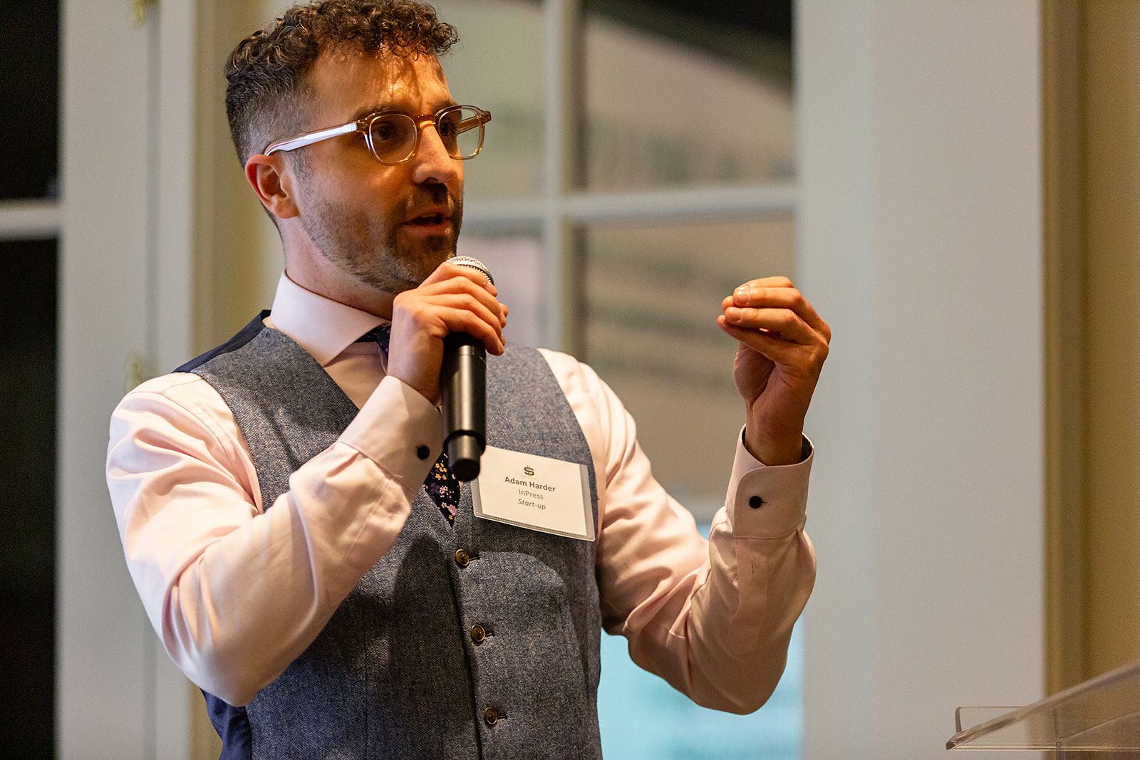 A man in glasses and a vest speaks into a microphone, gesturing with his left hand, while standing indoors near a window.