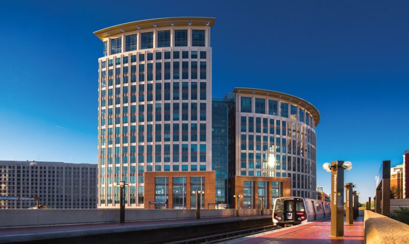 A modern office building with two curved towers stands behind a metro train arriving at an outdoor urban station under a clear blue sky.
