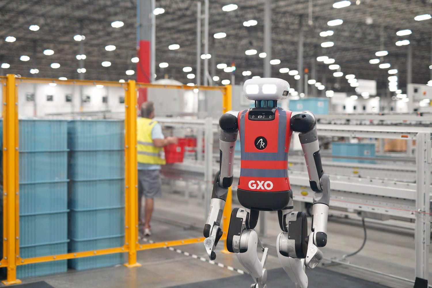 A humanoid robot wearing a GXO vest stands in a warehouse with conveyor belts, while a worker in a yellow vest operates in the background.