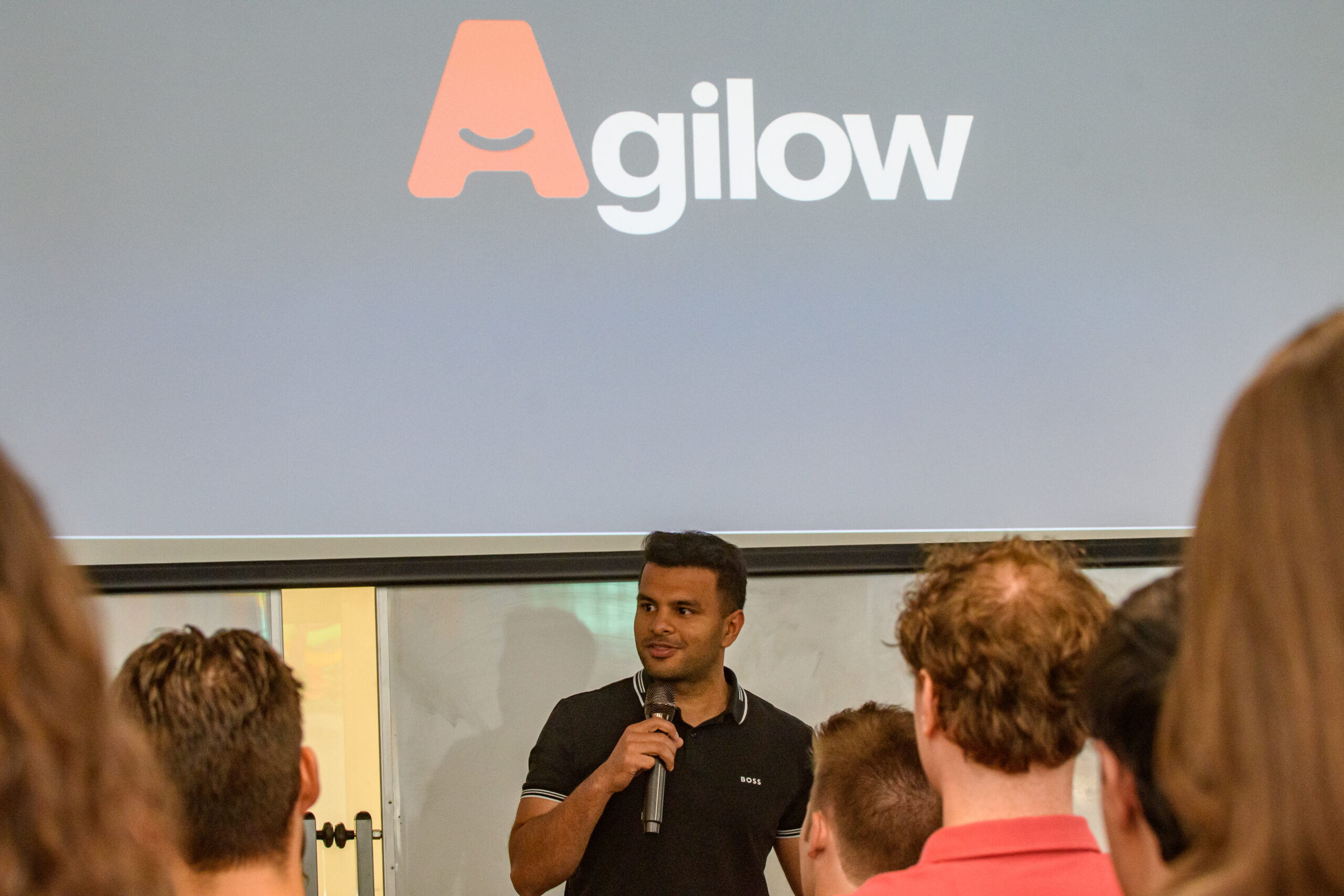A man holding a microphone speaks to an audience in front of a screen displaying the Agilow logo.