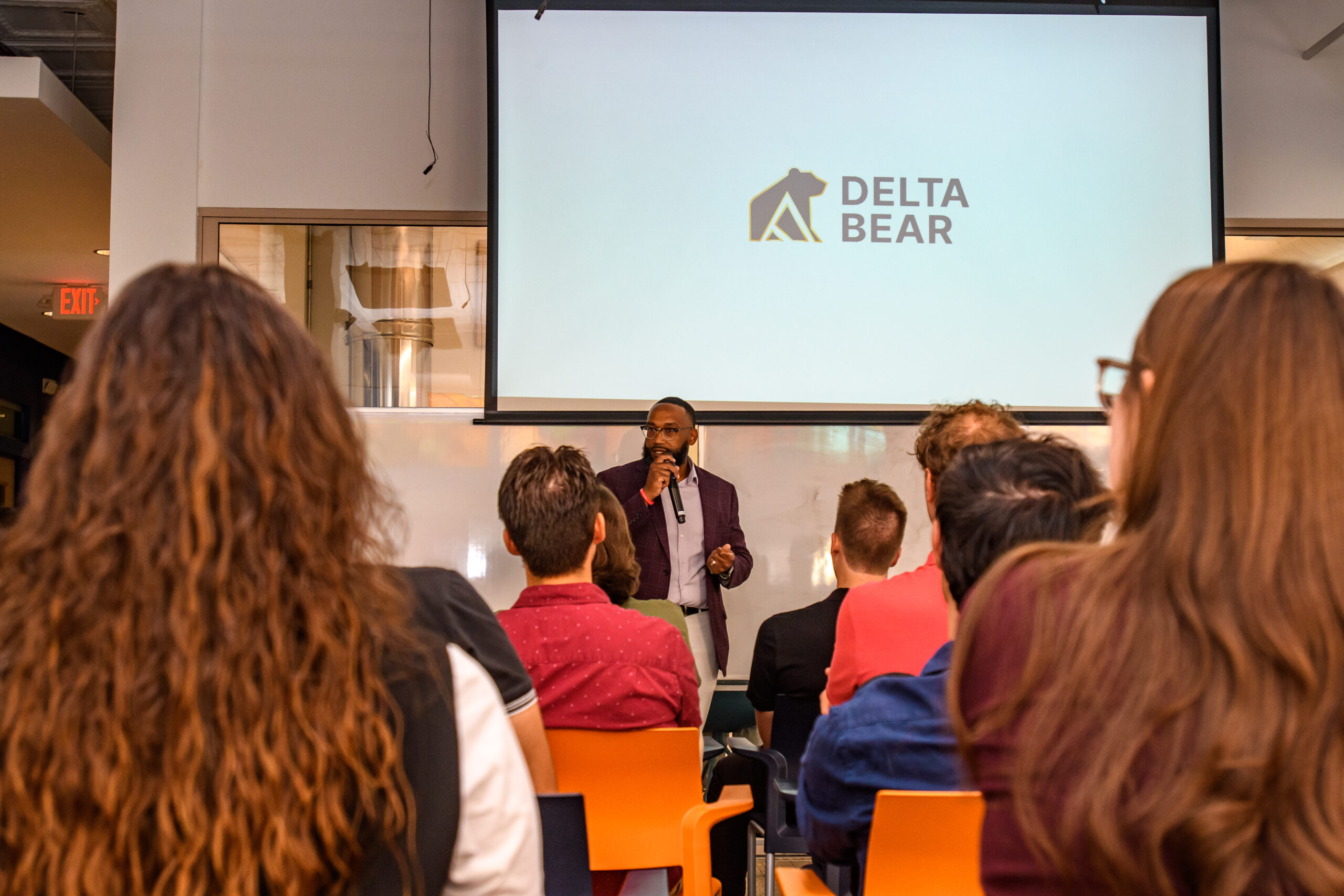 A man speaks at the front of a room to an audience, with a presentation slide displaying the Delta Bear logo behind him.