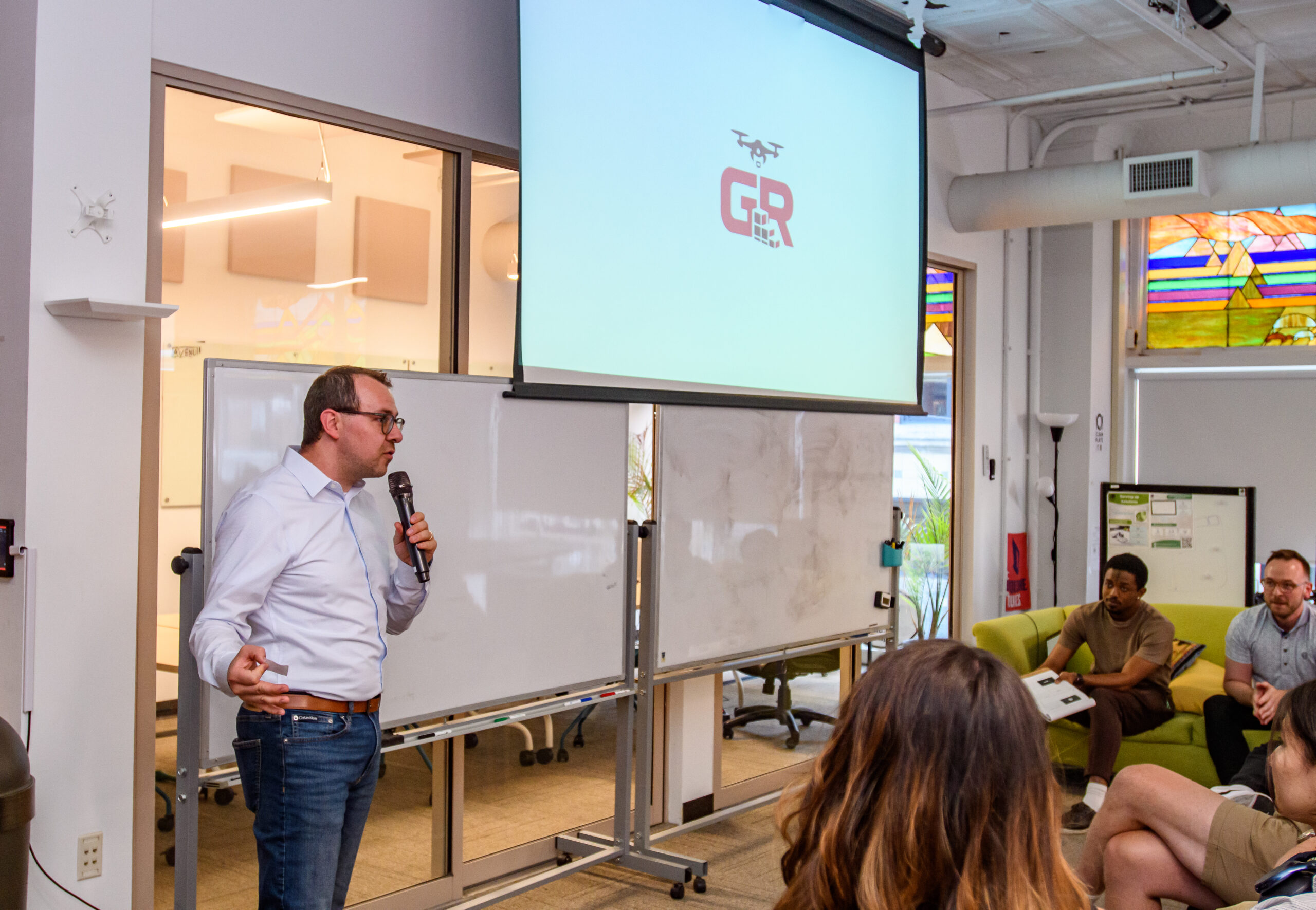 A man with a microphone presents in front of a screen displaying a logo to a seated audience in a modern office space.