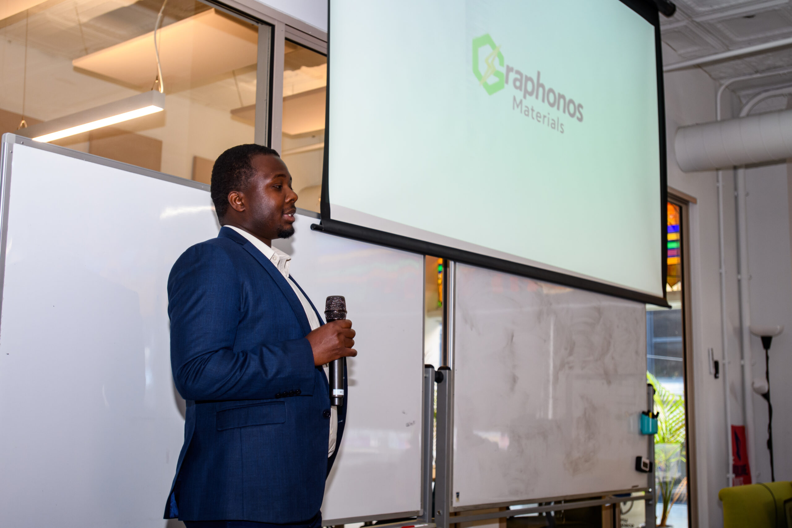 A man in a blue suit holds a microphone and presents in front of a screen displaying the "Graphonos Materials" logo in a conference room.