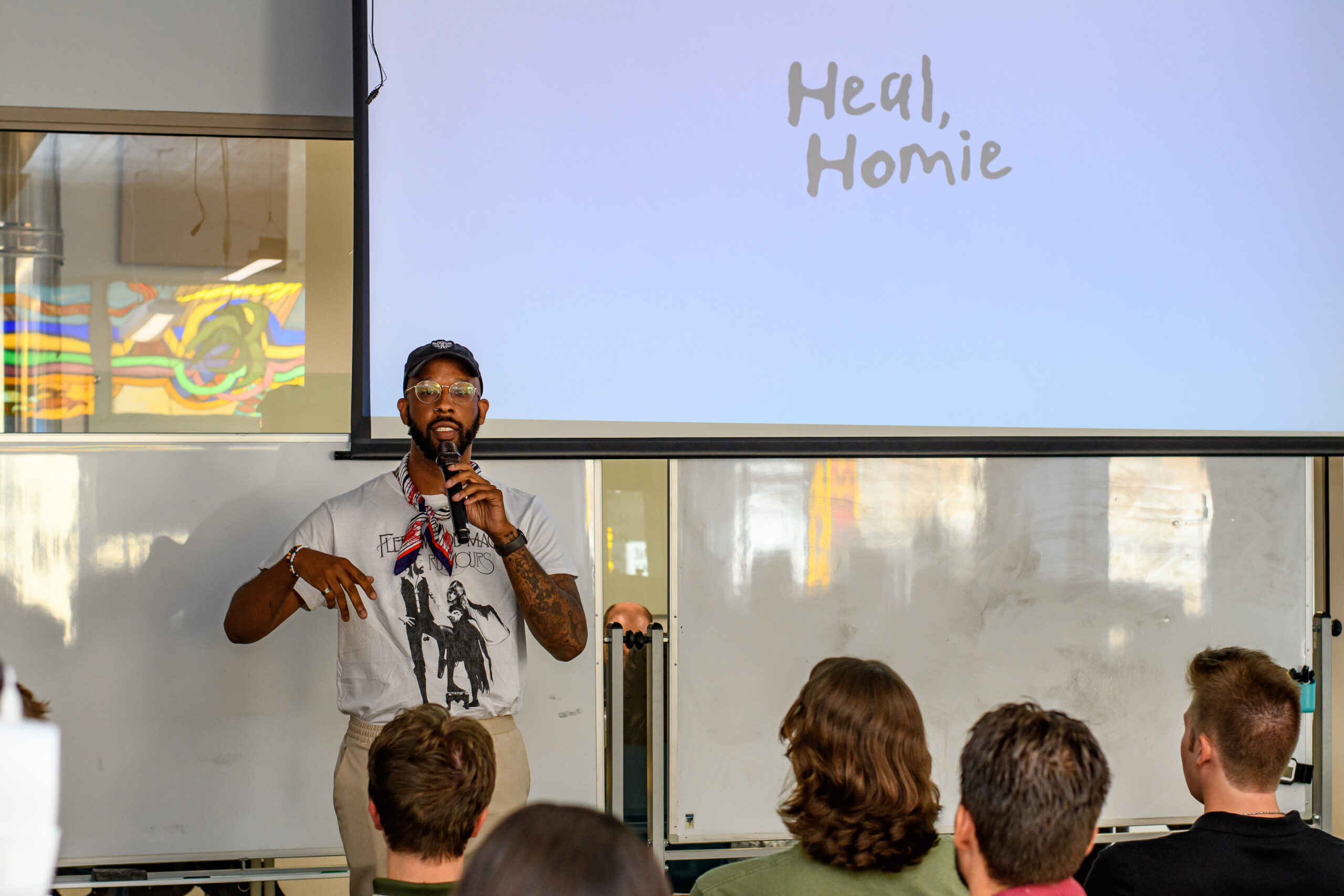 A speaker addresses an audience in a classroom, with a presentation slide behind him that reads "Heal, Homie.
