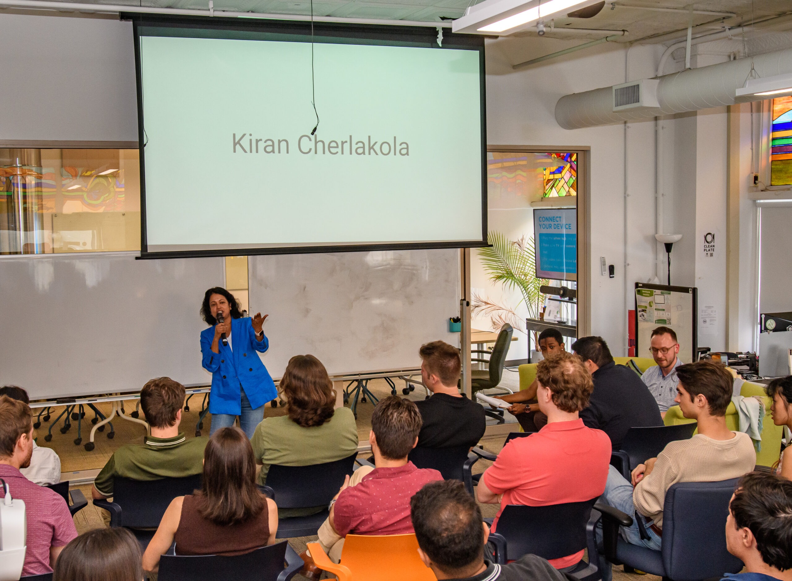A woman in a blue suit speaks to an audience in a classroom. A projector displays the name “Kiran Cherlakola” on the screen behind her.