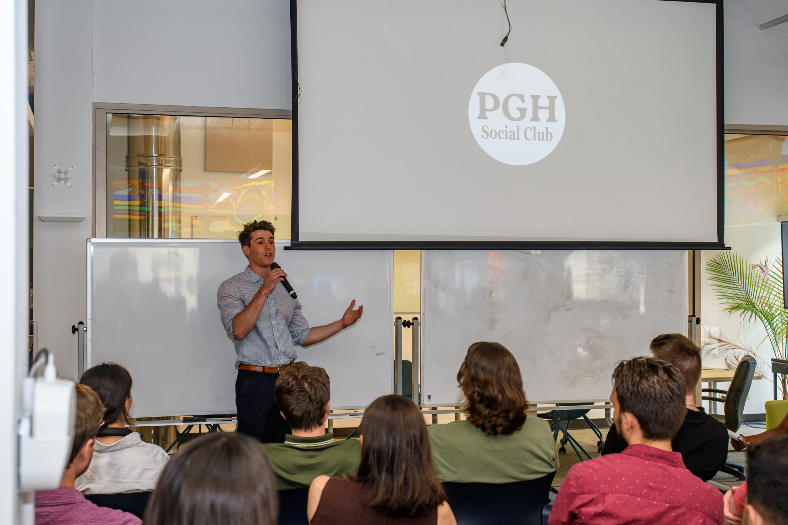A man stands and speaks into a microphone in front of an audience, with a projection screen displaying "PGH Social Club" behind him.