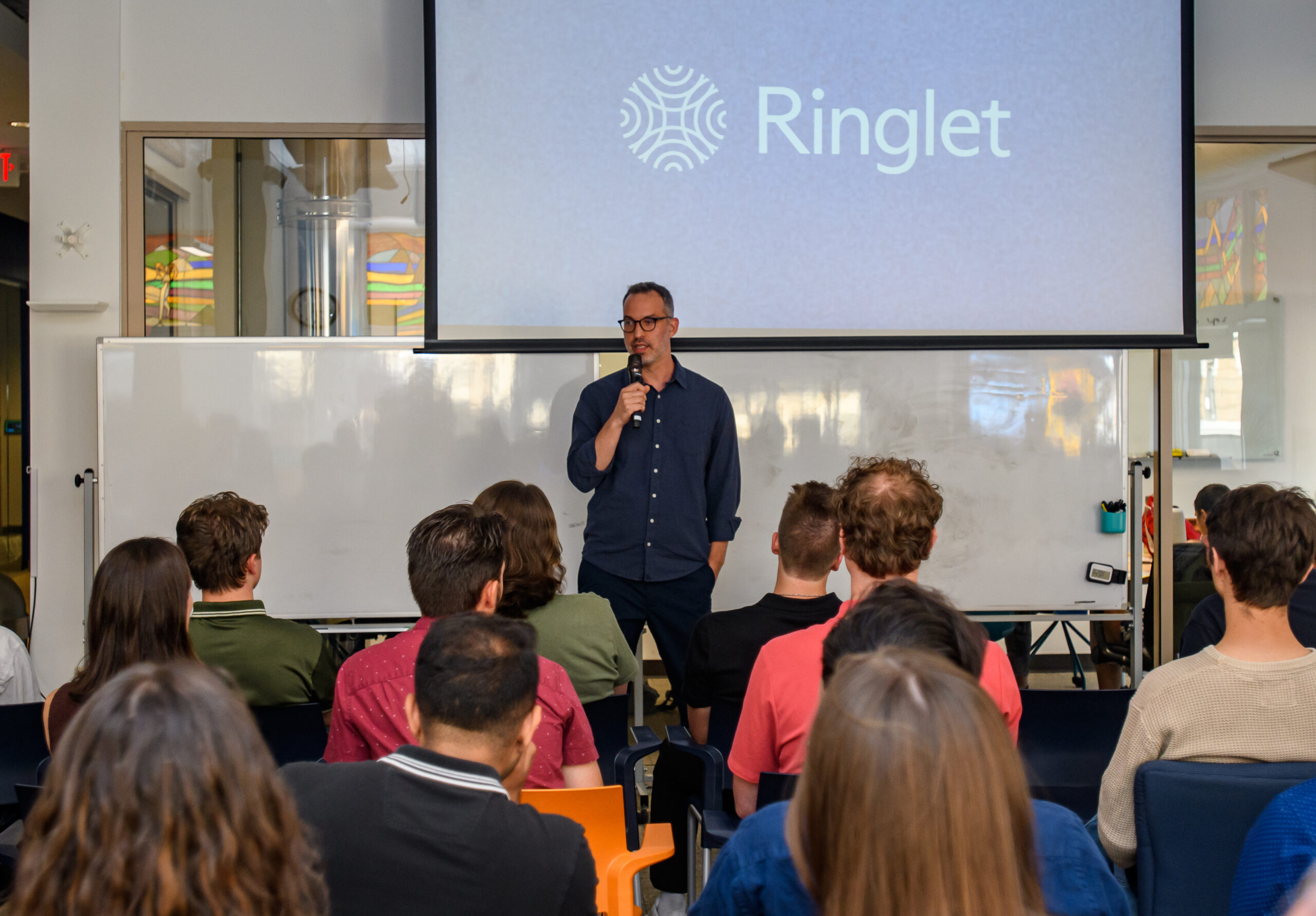 A man stands in front of a seated audience, speaking into a microphone, with a presentation slide displaying the word "Ringlet" behind him.