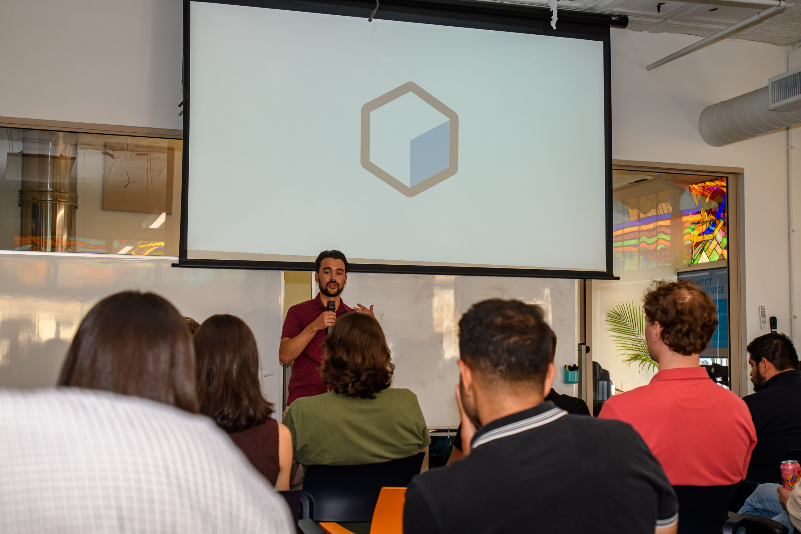 A man stands and speaks in front of an audience in a classroom setting, with a large screen behind him displaying a hexagonal logo.