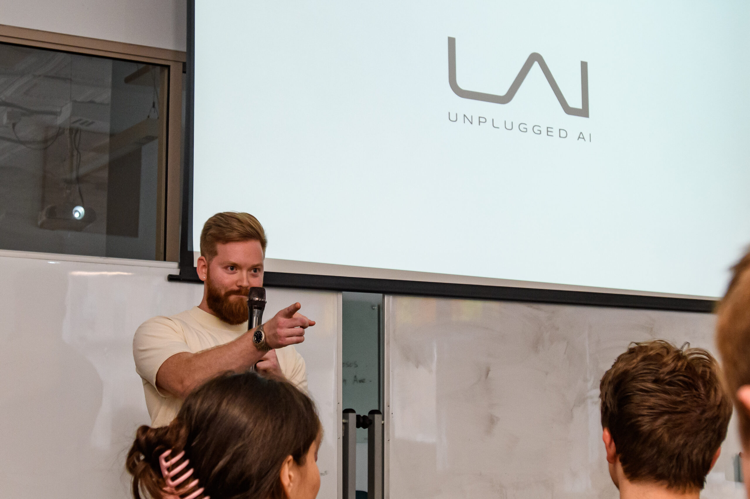 A man stands in front of a projected screen with the "Unplugged AI" logo, addressing an audience in a classroom or seminar setting.