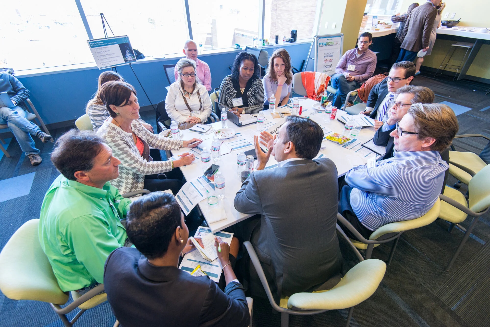 A group of people sit around a round table in an office setting, engaged in discussion with papers, notebooks, and drinks on the table.