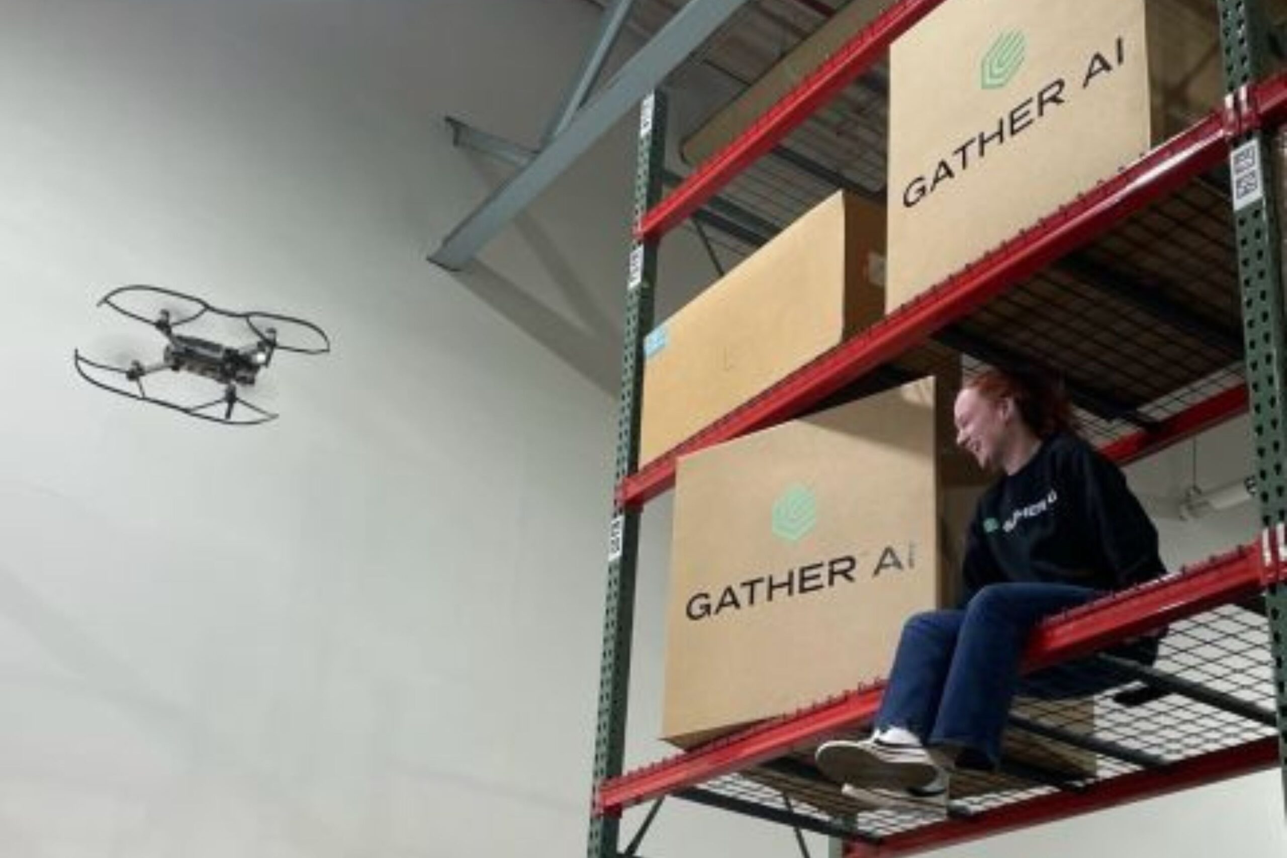 A person sits on a warehouse shelf next to large boxes labeled "Gather AI" while a drone flies nearby inside the warehouse.