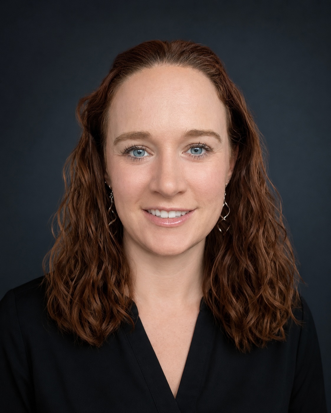 Woman with wavy auburn hair, light skin, and blue eyes, wearing a black top and silver earrings, posing against a dark background.