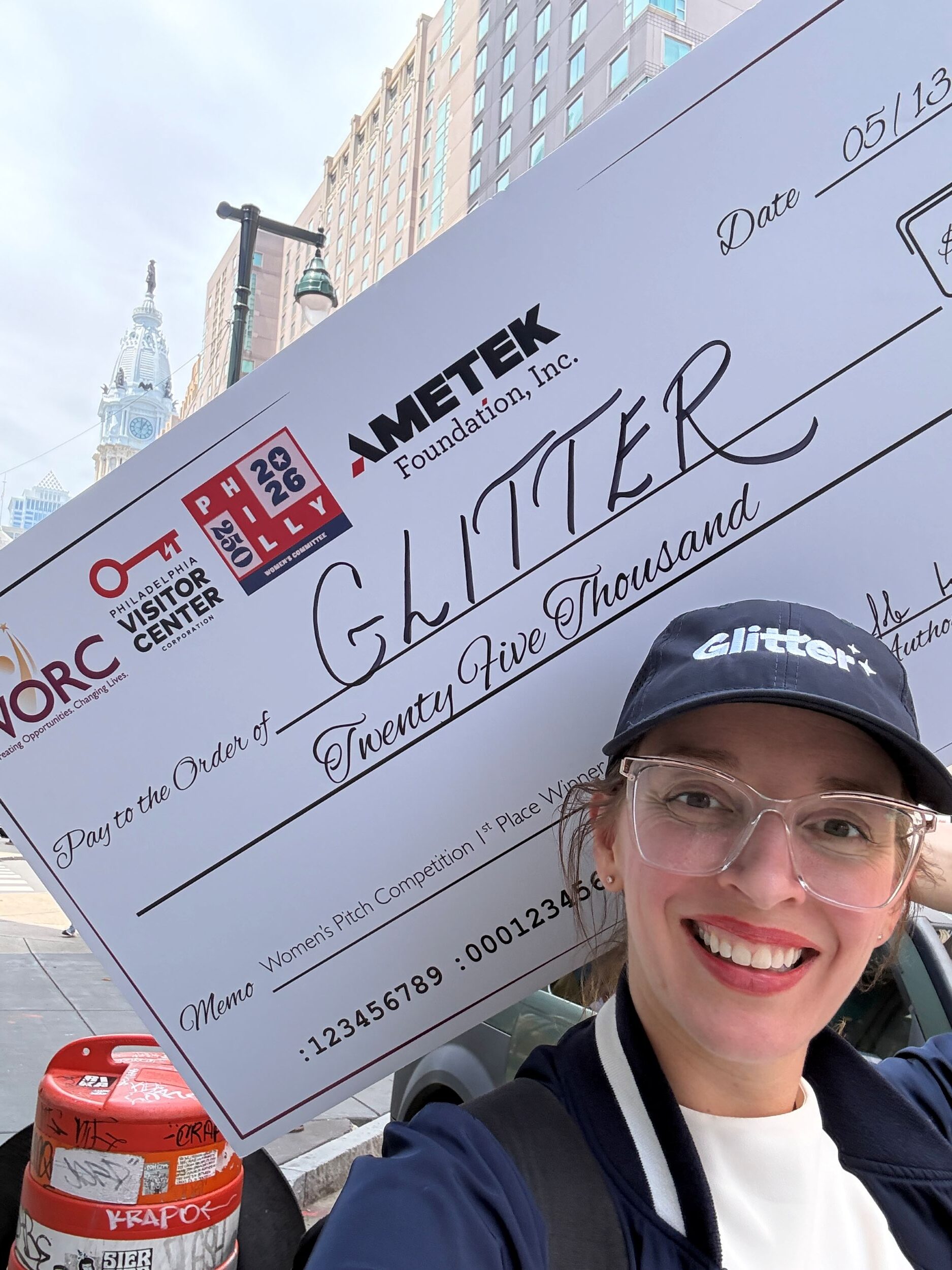 A person wearing a “Glitter” hat and glasses smiles, holding a large $25,000 check labeled “Glitter” in a city setting with buildings visible in the background.