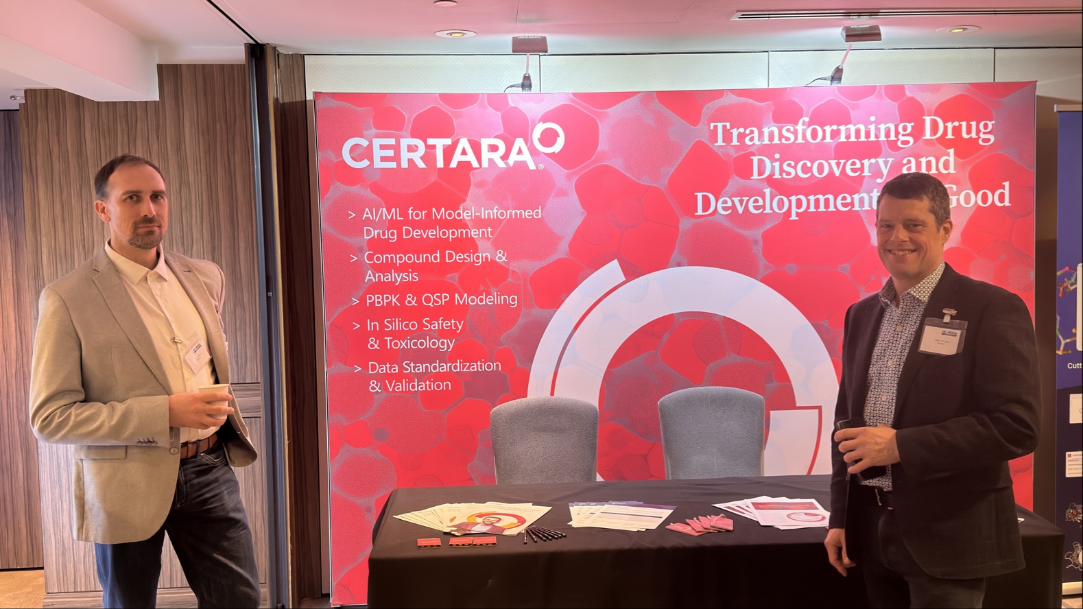 Two men stand at a Certara booth with a red backdrop displaying text about drug discovery and development. A table in front holds papers and promotional items.
