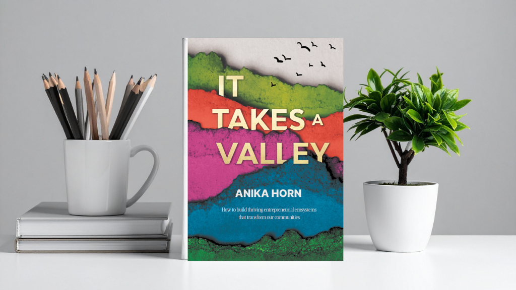 A book titled "It Takes a Valley" by Anika Horn stands on a desk between a potted plant and a cup of pencils, with stacked books underneath the cup.