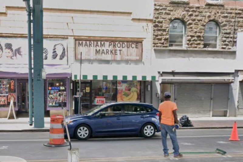 A person in an orange shirt stands on a city sidewalk near a traffic cone and a blue car parked in front of Mariam Produce Market.