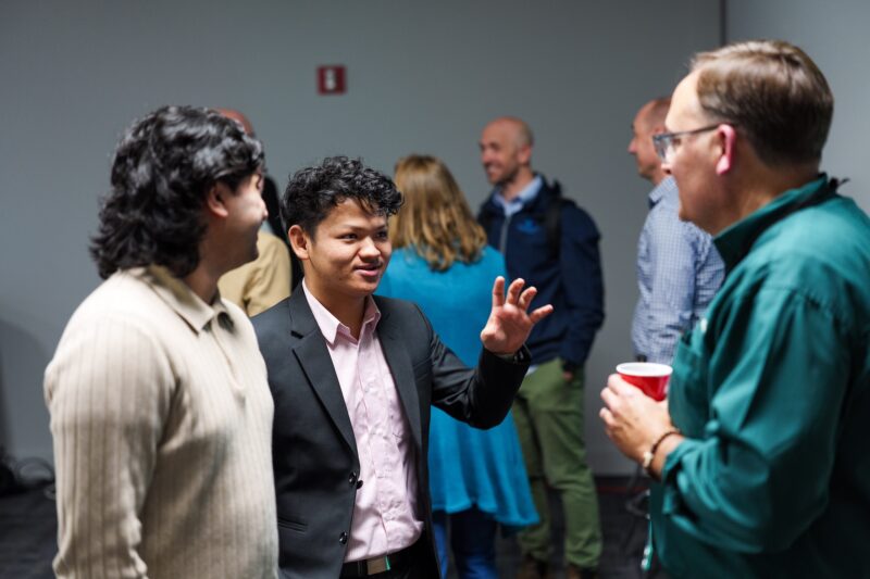 A group of people converses in a room; one man in a suit gestures while talking, and others listen and hold drinks.