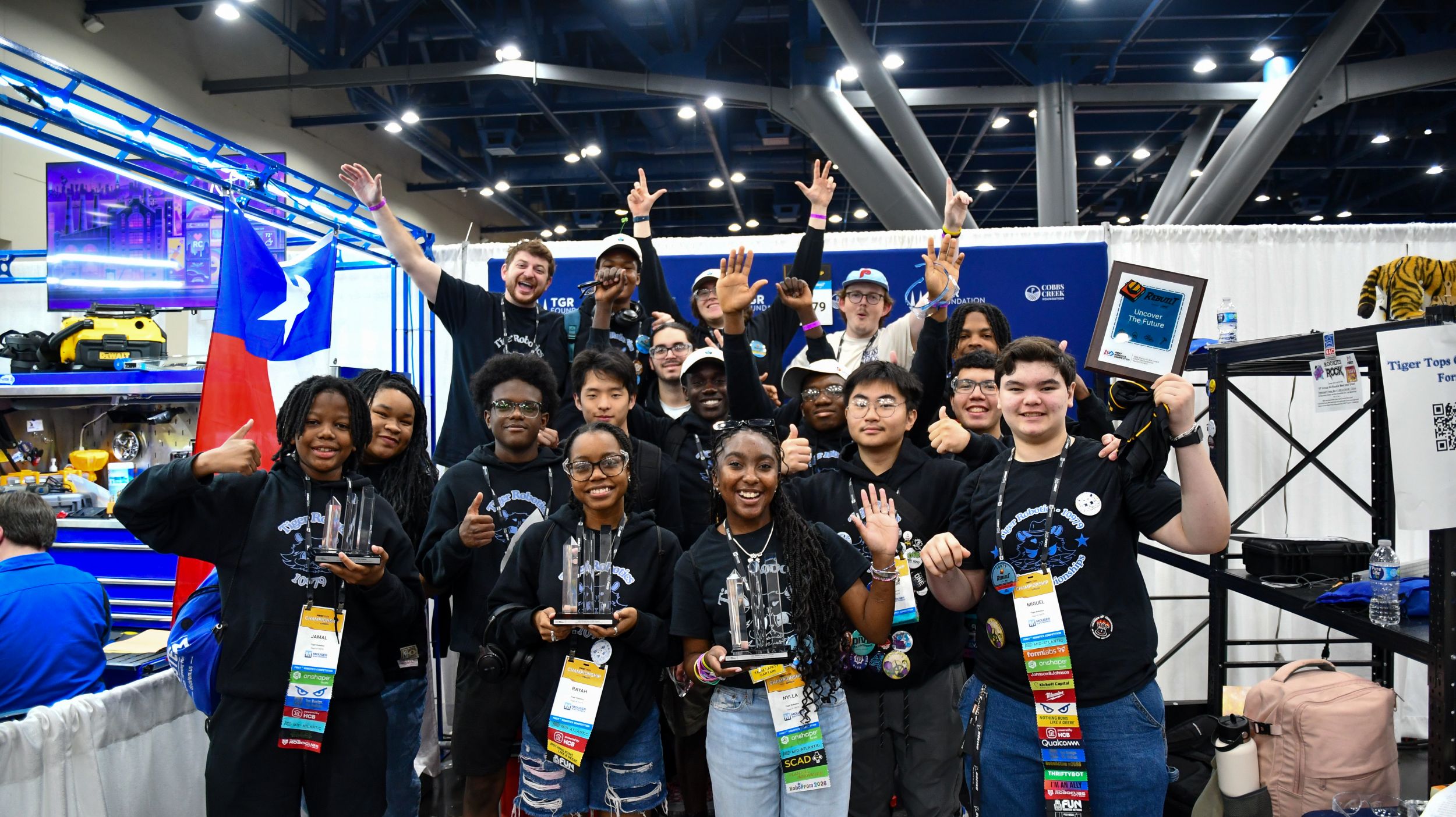A group of students in matching shirts pose and smile with trophies and medals at a robotics competition, standing in front of a Texas flag and team equipment.